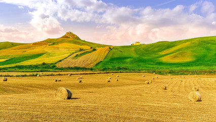 Obraz premium Scenic view at picturesque golden sunset valley in a green shiny field with hay stacks, bright cloudy sky , golden sun rays and beautiful hills on background, summer valley landscape