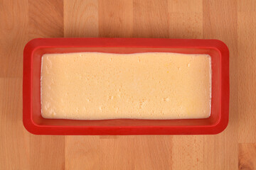 An overhead shot of a red rectangular silicone loaf pan filled with a pale yellow, unbaked lemon cake batter on a light wooden surface
