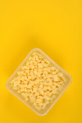 An overhead shot shows a clear, square container filled with yellow cheese cubes, diagonally placed on a vibrant yellow background with ample copy space