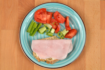 An overhead shot of a light blue plate holding an open-faced sandwich with ham and vegetables, including sliced tomatoes and celery, set on a wooden surface