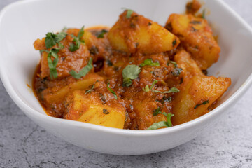 Aloo Curry – Traditional Indian potato curry made with tender potatoes simmered in a spiced tomato gravy with herbs and aromatic seasonings, served in a white bowl on a textured stone background