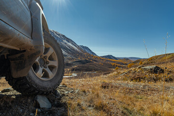 An off-road tire stands on yellow grass during autumn, overlooking a river and snow-capped mountains. An off-road wheel against the backdrop of mountains. A tire against the backdrop of mountains.