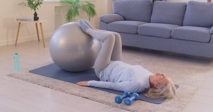 Senior lady having fitness workout with sports equipment. Old woman lying on yoga mat in supine position with feet placed on stability fit ball does knee bends for being in good shape and joint health