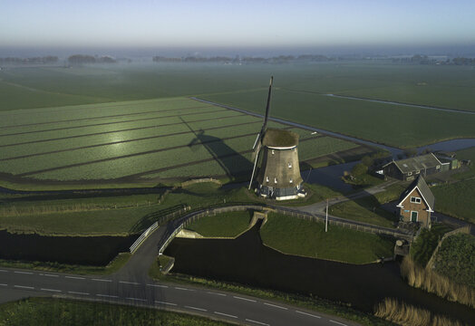 Aerial view of a classic windmill standing tall amidst the flat, verdant fields, casting a long shadow under the clear sky, a serene tableau of rural charm, Groningen, Netherlands.