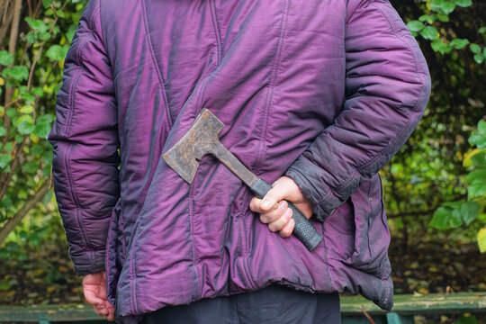 A man in a purple jacket holds a gray black axe in his hand behind his back on an autumn street
