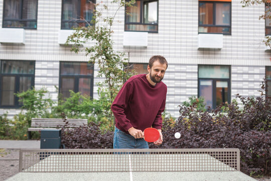Man in a burgundy sweatshirt playing table tennis outdoors near a modern residential building, holding a red paddle and focusing on the game, symbolizing leisure, activity, and a healthy lifestyle - Powered by Adobe