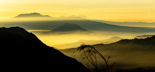 View of golden light blankets the misty valleys and silhouettes of distant mountains, a serene landscape that whispers ancient tales, Bromo, East Java, Indonesia.