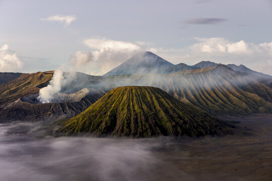 View of volcanic peaks pierce through a sea of ethereal mist, plumes of smoke rising, creating a dramatic, otherworldly landscape, Bromo, East Java, Indonesia.