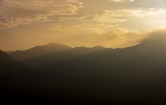 View of golden light illuminating the layered mountain range under a soft, cloud-strewn sky, casting long shadows across the peaks and valleys, Bromo, East Java, Indonesia.