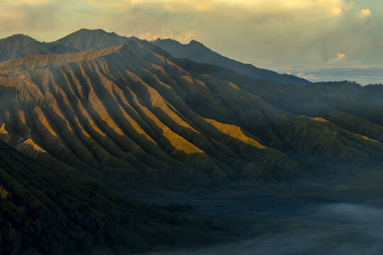 View of a mountain range, with sunlight casting golden rays across the ridges, set against a backdrop of a soft sky, Bromo, East Java, Indonesia.