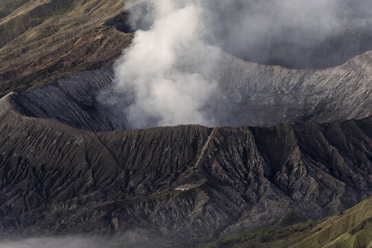View of a volcanic crater hisses with geothermal activity, its dark, textured slopes contrasting against the ethereal smoke, Bromo, East Java, Indonesia.