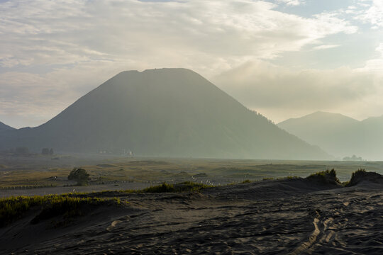 View of the majestic mountain peak rising through a misty haze over a textured landscape under a partially cloudy sky, Bromo, East Java, Indonesia.