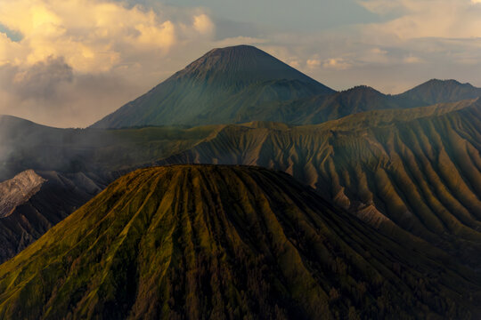 View of majestic volcanic peaks with rugged textures and wisps of smoke against a backdrop of warm sunlight and dramatic sky, Bromo, East Java, Indonesia.