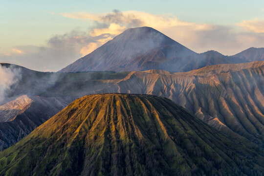 View of the dramatic volcanic landscape with sharp ridges and a dominant cone bathed in golden light against a soft sky, Bromo, East Java, Indonesia.