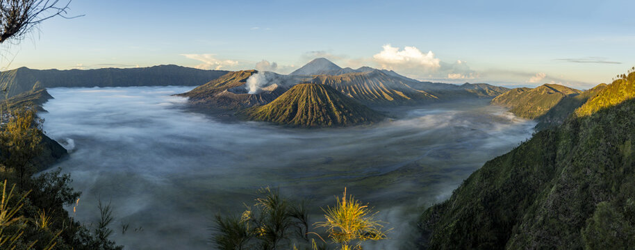 View of mist-shrouded volcanic landscape with smoking peaks piercing through the dense fog under a bright sky, Bromo, East Java, Indonesia.