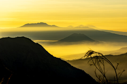 View of the silhouette of rugged mountains piercing through golden mist, bathed in the warm glow of sunrise, a scene of ethereal beauty, Bromo, East Java, Indonesia.