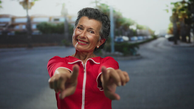 Senior woman with grey hair and red outfit smiling and pointing at the camera on a city street with blurred urban background and palm trees.