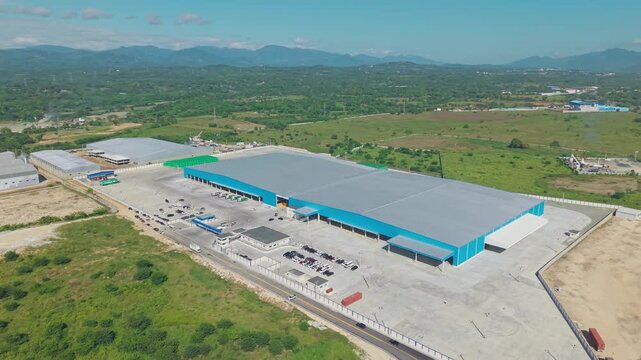 Aerial orbit shot of large Logistics Transformation Hub of Dominican National Brewery, Hato Nuevo, Santo Domingo. Sunny day with mountains in distance.