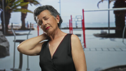 Senior woman with grey hair wearing a sleeveless dress, touching her neck while standing thoughtfully on a seaside promenade with blurred beach and fitness equipment in the background.
