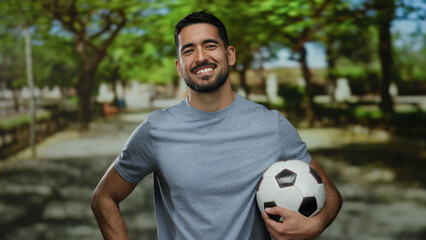 Young man holding soccer ball standing confidently in a lush green park with a bright smile on his face under the vibrant summer sunshine. © Krakenimages.com