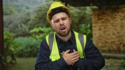 Young construction worker wearing yellow hardhat and hi vis vest clutching chest beside stone building in countryside; pain emergency.