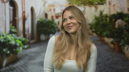 Blonde woman wearing white top touches hair in sunlit ancient cobblestone alley; serenity confidence.
