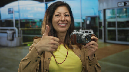 Middle age hispanic woman holds vintage camera and shows thumbs up at airport terminal outdoors; approval.