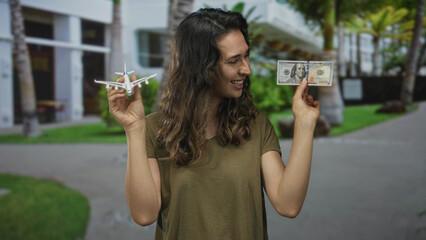 Woman smiling while holding airplane model and dollar bill on sunny busy street; travel planning...
