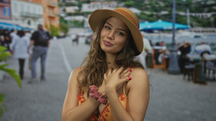 Young hispanic woman in sunhat and orange bikini makes finger heart gesture on street outside seaside restaurant; affection.