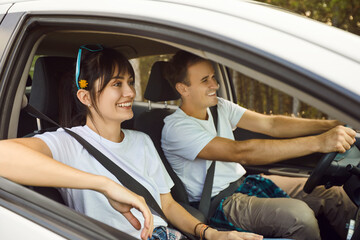 Happy couple of tourists drive rental car along rural road near summer forest. Weekend travel of smiling people enjoying carefree journey, discussing route and adventure, safe driving with seatbelts