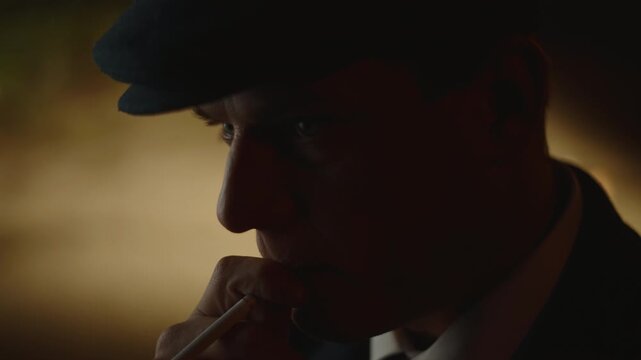 Mysterious mature man wearing a vintage flat cap and suit smoking a cigarette in a dark, moody room, with his face lit in silhouette creating a dramatic and pensive atmosphere