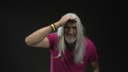 Man with long gray hair and beard holding hand to forehead shielding eyes in black studio wearing magenta shirt and bracelets; curiosity exploration.