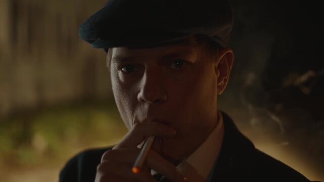 Serious gangster in vintage attire lighting and smoking a cigarette in a dark, atmospheric setting, looking intently at the camera while exhaling a plume of smoke from his mouth