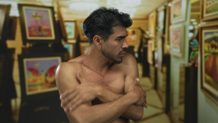 Man wraps arms across bare chest inside gallery building among colorful paintings while shivering with cold; vulnerability.