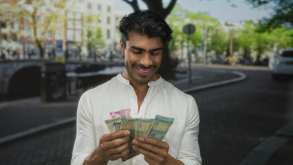 Man standing on a crowded street counting indian rupee banknotes with his hands as people pass by; contentment.