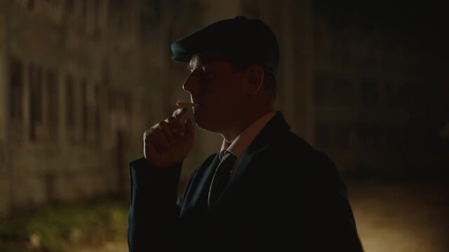 Handsome vintage-style gangster wearing a flat cap and suit thoughtfully smoking a cigarette at night on a dark, atmospheric street, with smoke billowing from his mouth