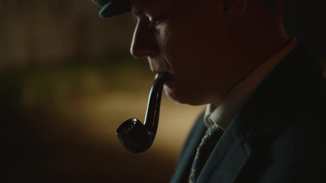 Serious man wearing a retro-style suit and flat cap lighting up a traditional wooden tobacco pipe with a match and exhaling smoke in a dimly lit, atmospheric room during the evening