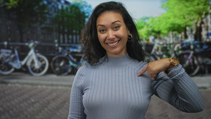 Young hispanic woman pointing finger to her shoulder on an amsterdam canal street with bicycles...