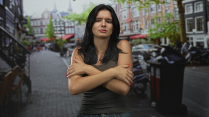 Young hispanic woman crossing arms on shoulders in wet cobblestone street after rain; quiet inner vulnerability.