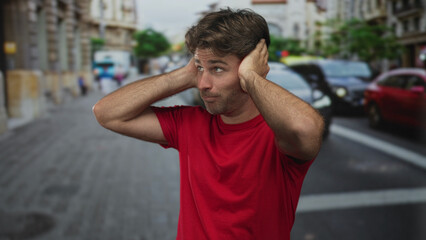 Young hispanic man in red t shirt covering ears with hands amid busy downtown street traffic and...