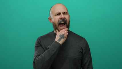 Man with a beard yawning against a solid green background, showcasing a bald head and grey sweater, exuding a handsome, tired expression in a minimalist setting.