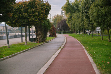 Empty curved pedestrian and bicycle paths run parallel through a green urban park with trees and benches under an overcast sky, beside a calm river waterfront for a peaceful stroll.
