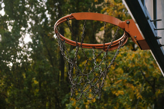 Close-up view of an outdoor basketball hoop with a metal chain net set against blurred autumn foliage, conveying rustic urban sports atmosphere and textured rim details in soft light.