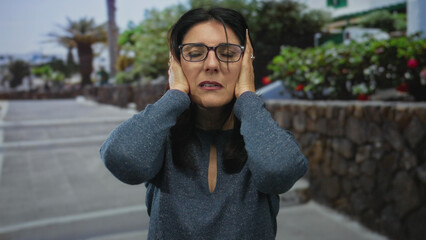 Fototapeta premium Middle age woman holds bare ears with hands on city street sidewalk against blurred stone wall and foliage; frustration.