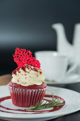 Delicious red velvet cupcake with cream cheese frosting and a touch of cocoa powder, elegantly presented. There's a decorative red element on top, alongside a sprig of rosemary.