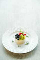 A beautifully presented fruit tart on a decorative white plate. The dessert is adorned with fresh blueberries, strawberries, and kiwi, complemented by sprigs of mint and rosemary