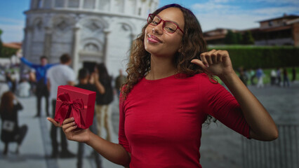 Fototapeta premium Woman in red shirt holds red gift box and points finger to cheek at building plaza near tower with tourists; confidence celebration travel.