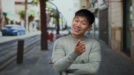 Fototapeta premium Young chinese man standing on a city street with buildings in the background, wearing a gray sweater and crossing his arms, showcasing a cheerful expression outdoors.