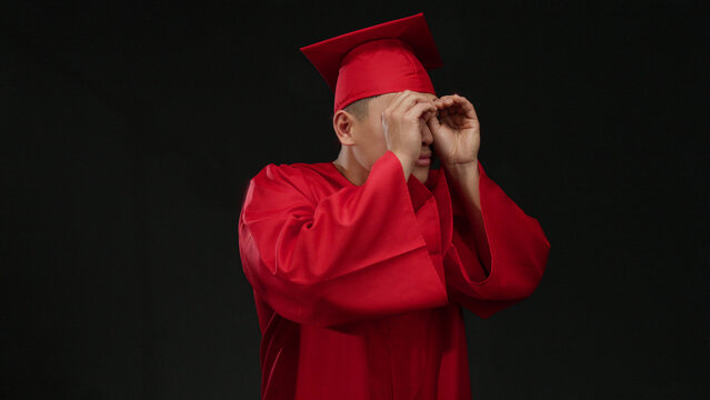 Young chinese man in red graduation cap and gown makes binocular gesture isolated on a black background, symbolizing curiosity and future vision.