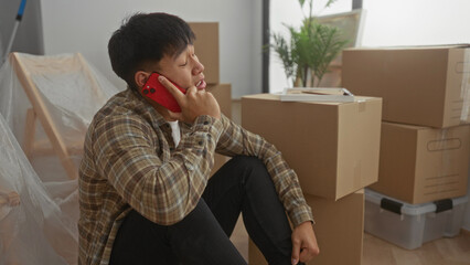 Young man sitting with phone surrounded by moving boxes in a new apartment living room, captured in...